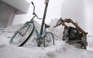 A bicycle is locked to a pole as a loader dumps snow onto a pile while clearing sidewalks in Boston, Thursday, Jan. 4, 2018. Forecasts for the Boston area call for about a foot of snow during the day and blizzard-like conditions. (AP Photo/Charles Krupa)